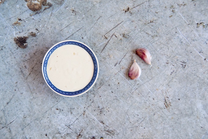 A bowl of garlic tahini dressing and two cloves of garlic viewed from above on a distressed metal background