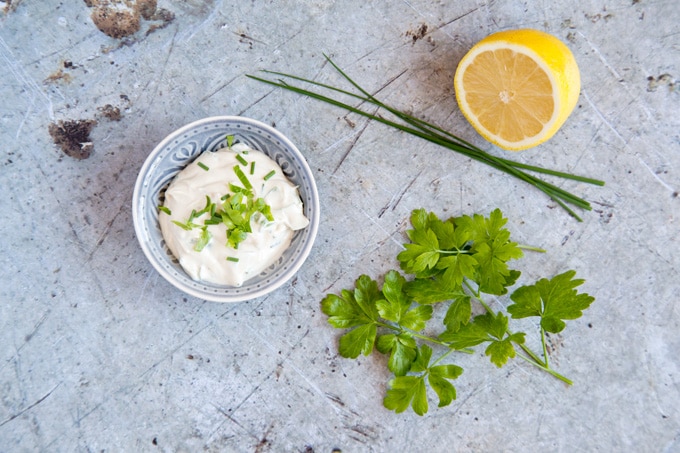 A bowl of lemon herb tahini dressing on a distressed metal background. Also in the photo is half a lemon, fresh chives and parsley