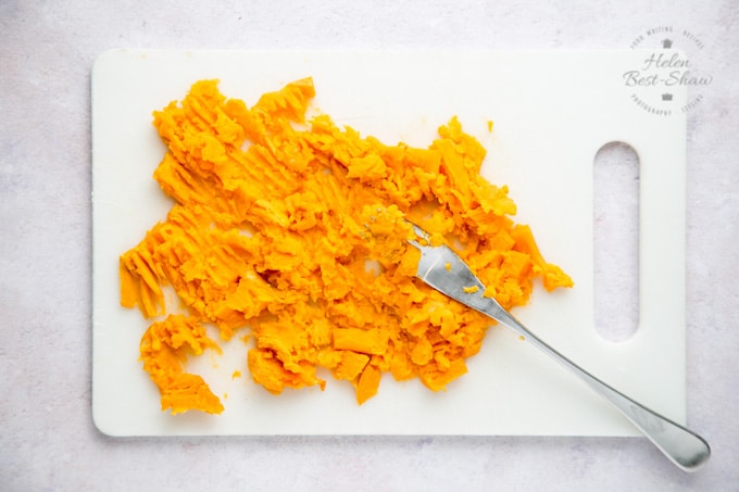 A chopping board covered with sweet potato which has been mashed with a fork