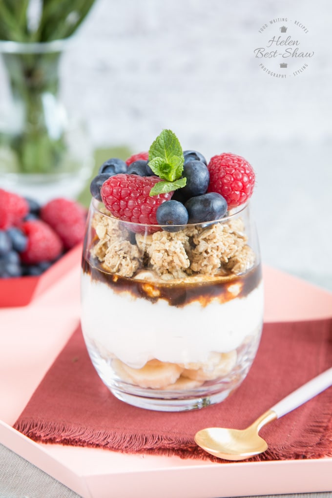 A healthy banoffee pie pot served in a glass consisting of layers of sliced banana, yogurt, soft brown sugar and topped with fresh berries garnished with a spring of mint. 