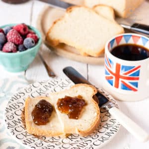 A plate with a slice of white yogurt bread spread with butter and jam. so shown is a loaf of bread, dish of berries and cup of coffee