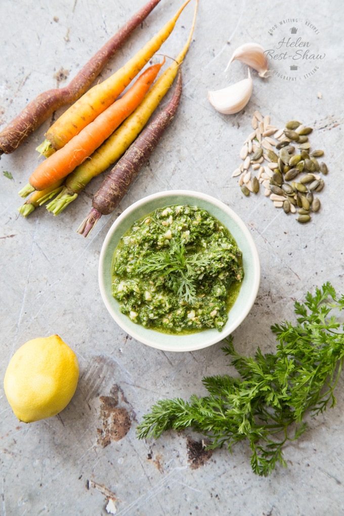A bowl of carrot top pesto on a grey work top surrounded by carrot tops, garlic, lemon and rainbow heirloom carrots viewed from above
