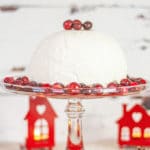 A glass cake stand with a domed white ice cream bombe on it, garnished with cranberries