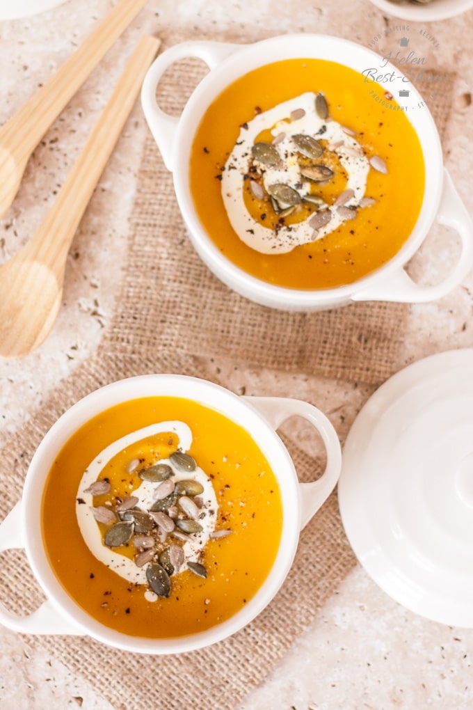 Two white bowls of golden orange pumpkin, carrot and sweet potato soup, drizzled with cream and toasted seeds viewed from above