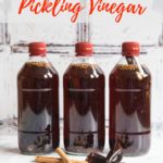 3 bottles of spiced vinegat for pickling standing on a marble background. A selection of spices including chillies, cinnamon sticks, cloves and cardamon pods are arranged in front. Text overlay readinf How to make pickling vinegar