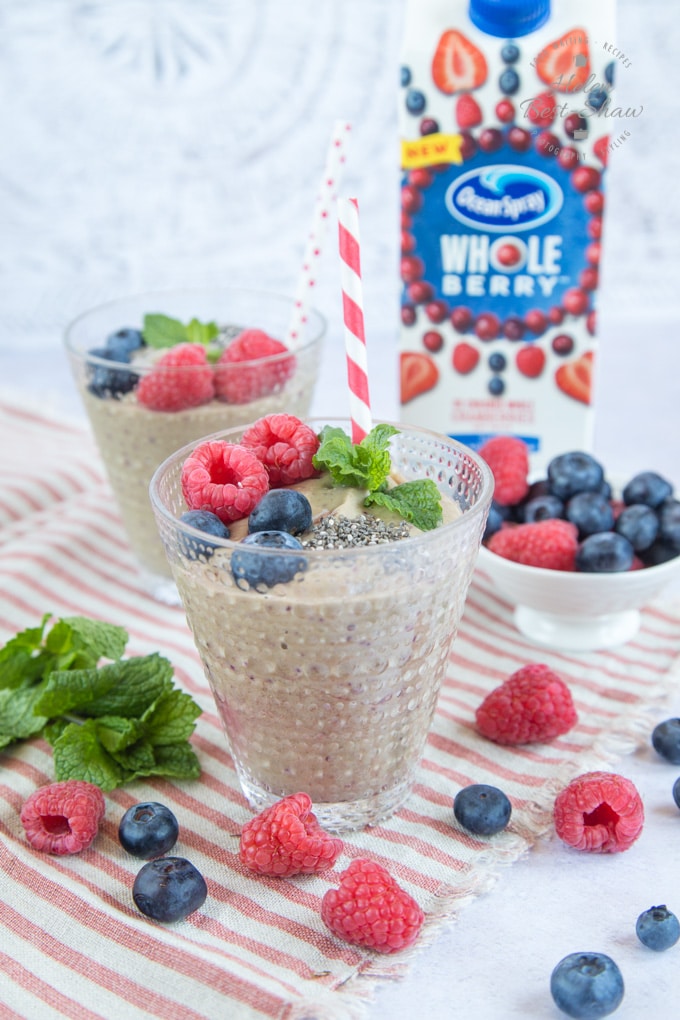 A pale green glass of cranberry smoothie with pea and avocado, topped with raspberries, blueberries, chia seeds and mint, ready to be enjoyed through a striped straw. In the background is a carton of Ocean Spray Whole Berry Cranberry Blueberry Raspberry Strawberry
