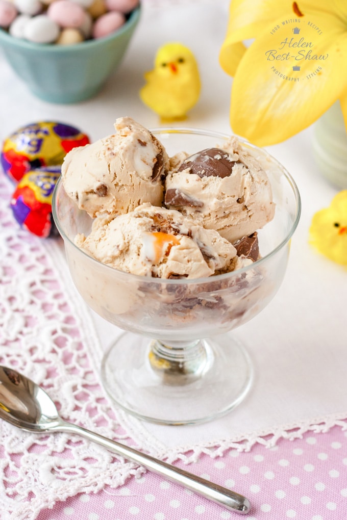 A glass sundae dish containing creme egg ice cream. The table is styled with a pink and white spotted cloth, Easter chocolates, chicks and yellow lilies 
