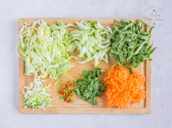 A wooden board covered with sliced vegetables: pak choi, carrots, spring onions, corainder and chili.