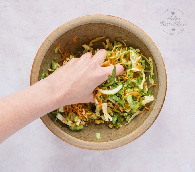 A salad bowl filled with Asian slaw; a hand with a handful of salad is tossing it to dress it.