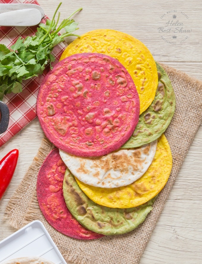 Pink, yellow, green and white coloured rotis on a hessian cloth. A sprig of coriander in the background.