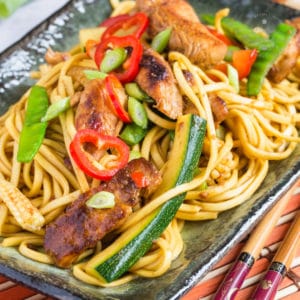 A close up on a rectangular plate of easy chicken chow mein. Noodles, chicken pieces, batons of courgette, mange tout peas and slices of chili can all be seen.