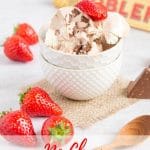 A white textured bowl full of Toblerone Ice Cream surrounded by strawberries and some chunks of chocolate. Also in the photo there is a wooden spoon and a packet of Toblerone Chocolate. Text overlay reads No Churn Toblerone Ice cream