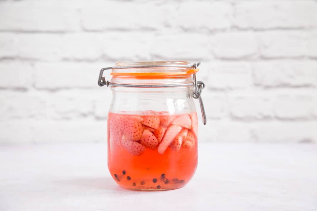 A mason jar of pale red black pepper and strawberry gin. The top half of the jar is full of floating strawberries. A few peppercorns can be seen sunk at the bottom. Thw strawberries take up much less space than they did when first placed in the jar.