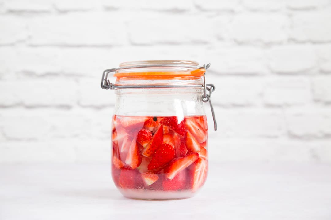 A side on shot of a mason jar just filled with black peppercorn, sliced strawberries and gin.