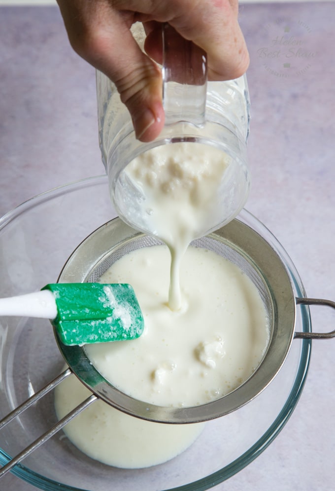 Kefir and grains in a jug are being poured into a sieve. The sieve sits in a glass bowl.