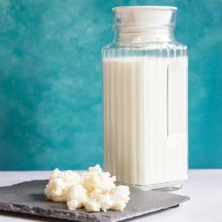 A jug of kefir against a blue background; in the foreground are some kefir grains on a slate.