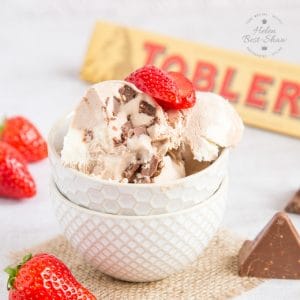A white textured bowl full of Toblerone Ice Cream surrounded by strawberries and some chunks of chocolate. Also in the photo there is a wooden spoon and a packet of Toblerone Chocolate