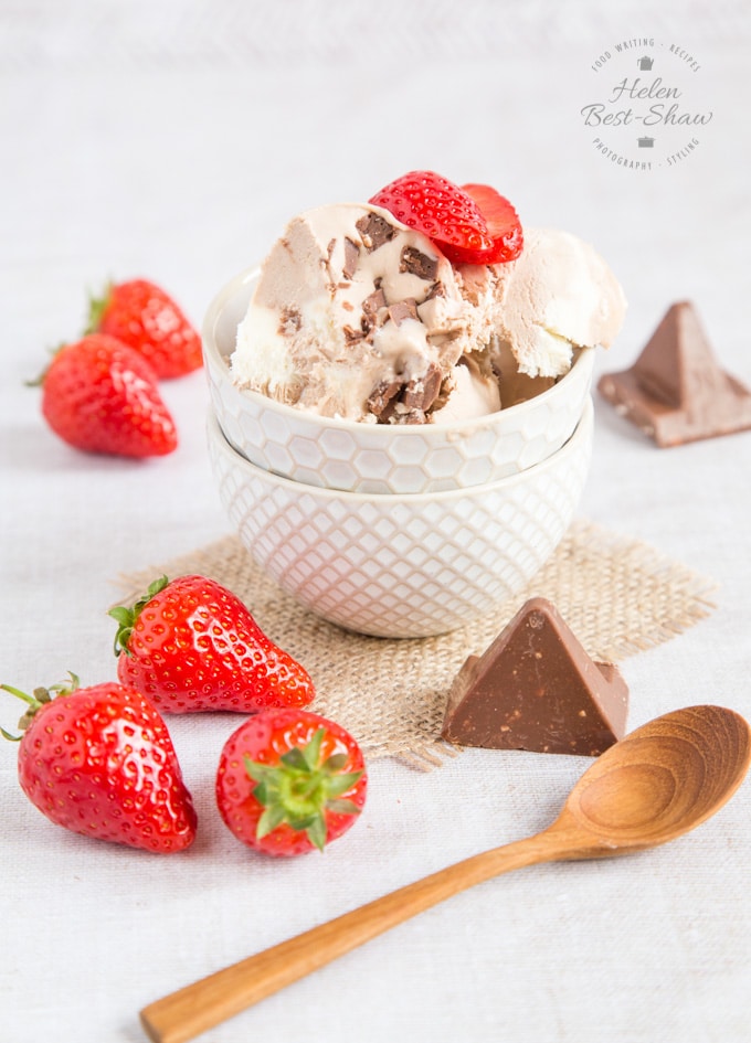 A bowl of no churn three ingredient Toblerone ice cream. Around the bowl are strawberries, triangular chunks of Toblerone, and a wooden teaspoon.