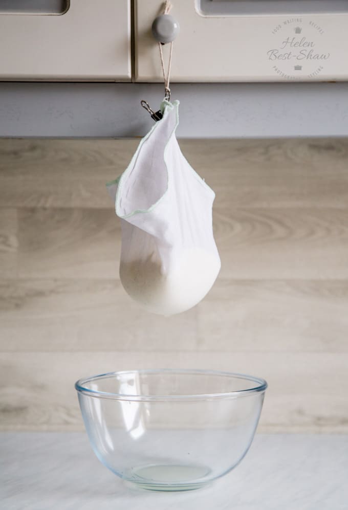 A muslin square filled with labneh, hanging from a kitchen cupboard. The whey is dripping into a glass bowl.