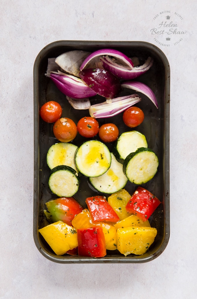 A top down view of Mediterranean vegetables - red onions, cherry tomatoes, courgettes, and red and yellow peppers - in a small roasting tray, covered with a dash of olive oil, ready to be cooked.