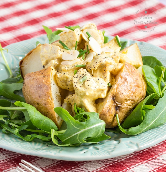 A baked potato topped with a thick helping of healthy coronation chicken, served on a bed of green salad. The dish is presented on a green plate, which is itself on a rad and white gingham table cloth.