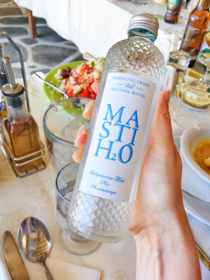A bottle of sparkling mastic water with a white label wtih blue writing. Held above a table of food