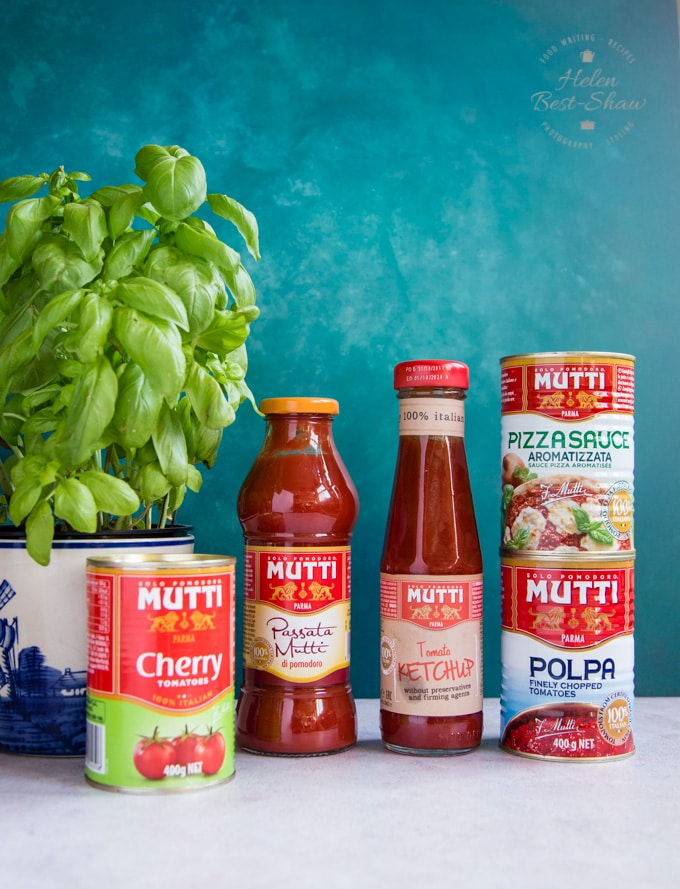 A selection of Mutti tomoato products - canned tomatoes, pizza sauce and passata. In the background there is a basil plant. 