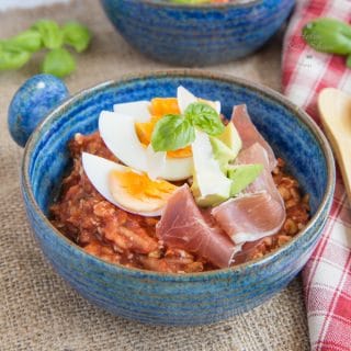 A close up on a bowl of savoury tomato overnight oats, topped with a quartered boiled egg and slices of air-dried ham.