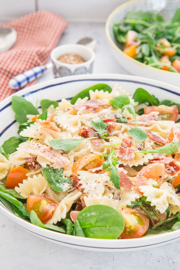 A shot of BLT Pasta salad on a blue rimmed plate, on green leaves. More green salad in the background, in a bowl. Also in the background are a white and red gingham cloth and a knife.