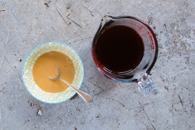 A glass jug of mixed soy sauce and white wine vinegar. Next to this is a small bowl containing some of this mixture to which some cornflour has been added, turning it into a opaque pale brown liquid; there's a teaspoon in the bowl. Both are on a rustic stone work surface.