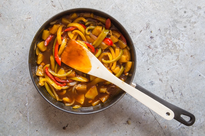 A frying pan of cooked peppers and pineapple chunks in a thickened sweet and sour sauce. The frying pan sits on a rustic stone work surface; a spatula coated with sauce sits in the pan.