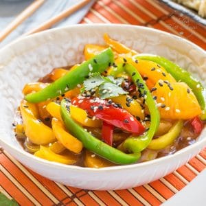 A close up on a bowl of peppers, coated with easy sweet and sour sauce and garnished with black and white sesame seeds. A dish of rice is out of focus in the background, along with a small bowl of more sweet and sour sauce. The main bowl is placed on a mat woven from wooden slats, alternating wider orange and thinner natural wood colours.