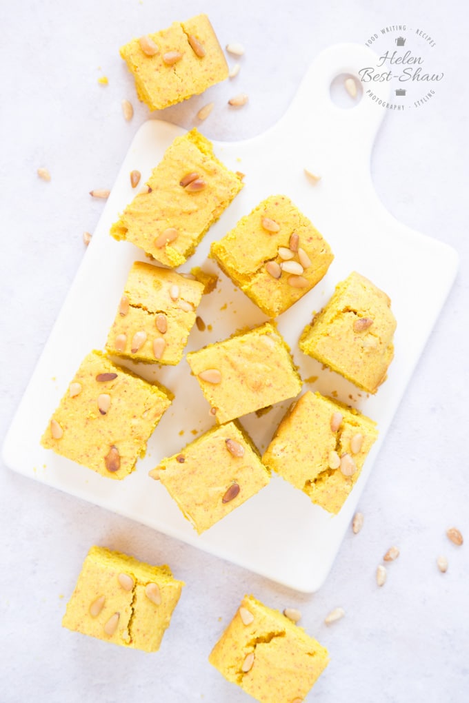 A top down view of slices of yellow sfoof turmeric cake on a porcelain board, with more slices on the grey work surface. Pine nuts are scattered about.