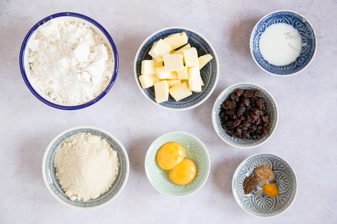 A shot of the ingredients for soul cakes: flour, sugar, butter, milk, raisins, egg yolks and spices, all arranged in seven small dishes laid out on a grey worksurface.