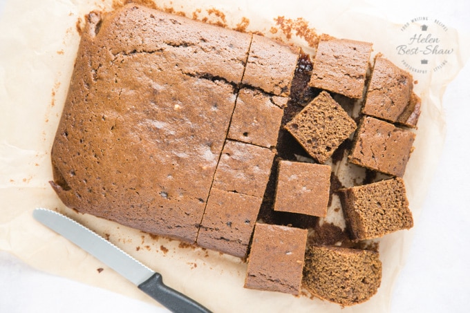 A picture from above of a baked rich brown rectangular easy vegan ginger cake sitting on greaseproof paper. Half the cake has been cut into squares. There’s a small black handled knife next to the cake.
