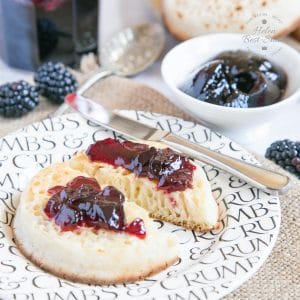 A close up of a crumpet spread with butter and blackberry jelly, on a sideplate with a knife. A small dish of more jelly is in the background. The plate sits on a rustic hessian cloth.