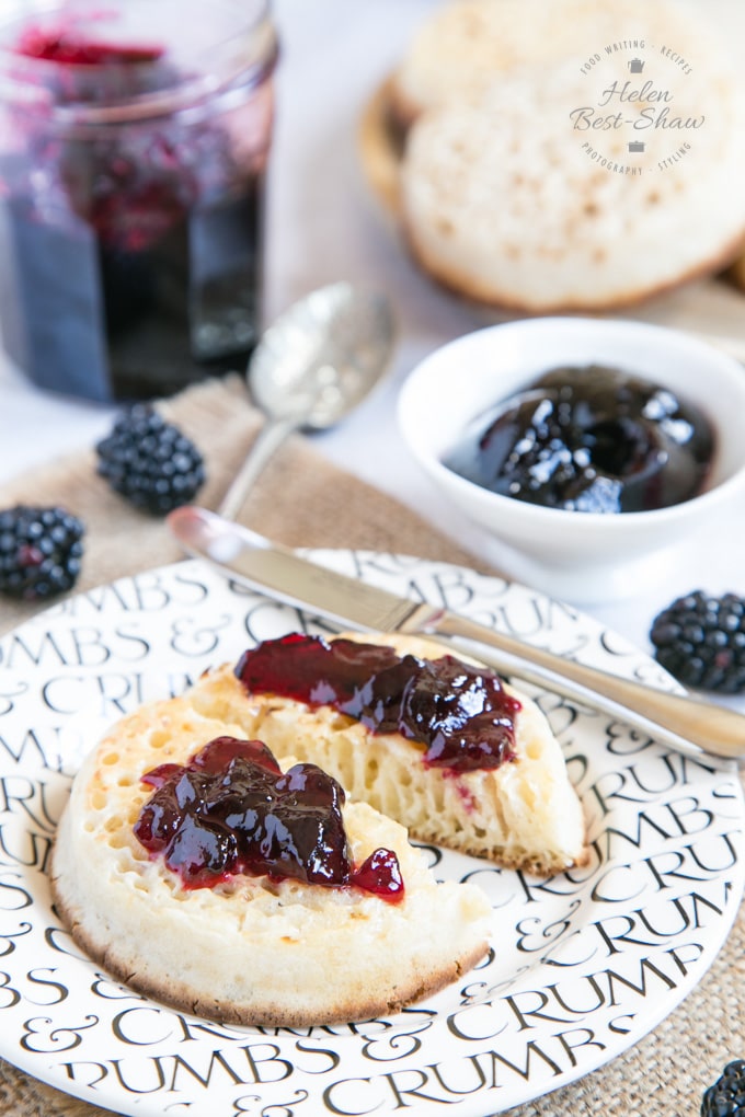 A close up of a toasted crumpet spread with butter and one pot blackberry jelly. The pot of jelly is out of focus in the background, along with more crumpets, a small dish of blackberry jelly and some individual blackberries.