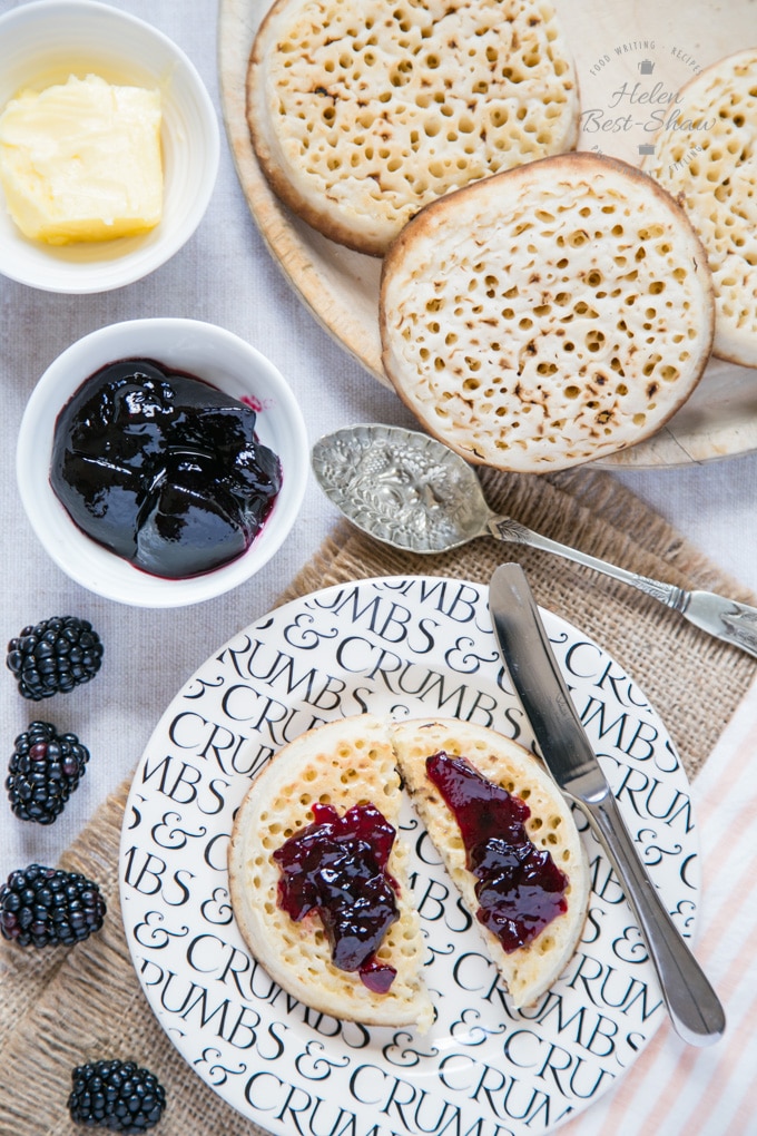 A sideplate with a toasted crumpet spread with butter and blackberry jelly. Three more crumpets are on a wooden breadboard at the top of the picture. Small dishes of butter and blackberry jelly are to the left. Individual blackberries are on the grey worktop.