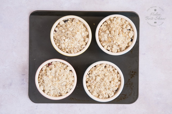 A top down shot of four individual apple and mixed berry crumbles ready for the oven. They are on a small grey baking tray, which is sitting on a grey worksurface. 