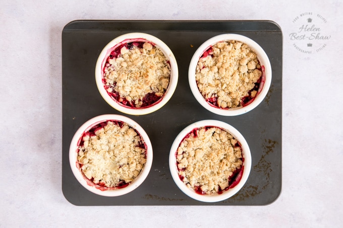 Cooked individual apple and mixed berry crumbles. The edges of the crumble are stained red from the berries underneath. The four pots are sitting on a grey baking tray, which itself is sitting on a pale grey worksurface. 