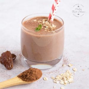 A close up of a glass of chocolate and date smoothie, with dates, cocoa powder and oats next to the glass.