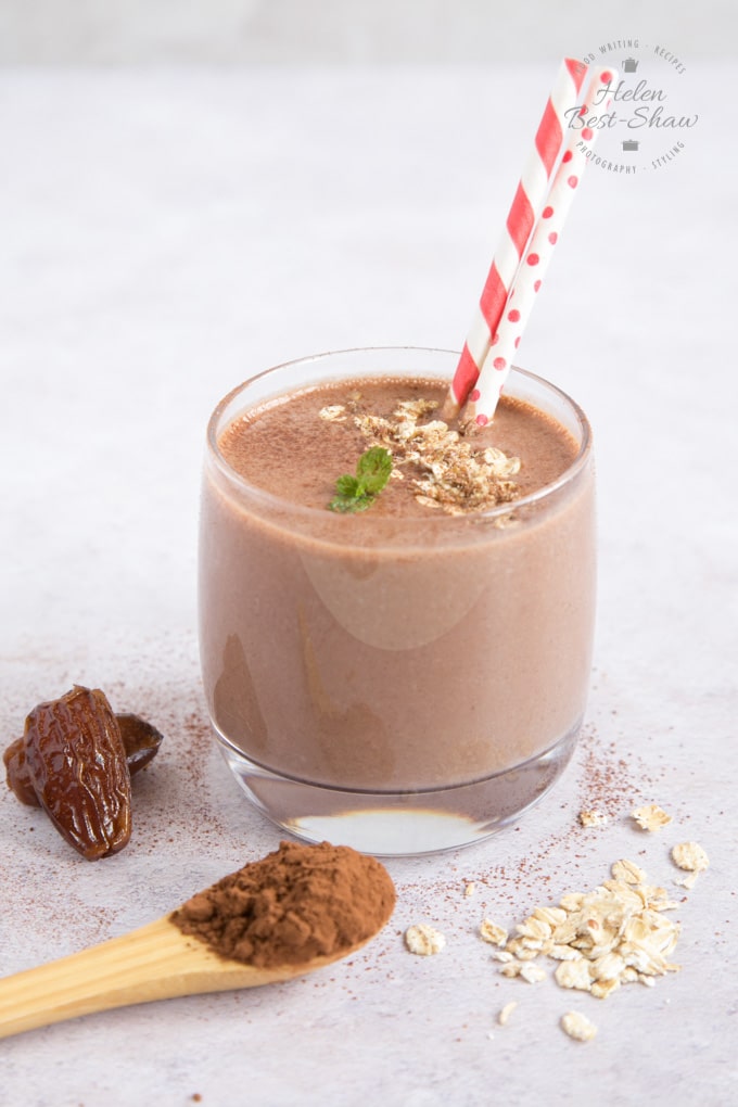 A photo of a glass of chocolate and date smoothie, in which sits two straws. The drink is topped with some oats and a small sprig of mint. Next to the glass are two dates and a small wooden spoon of cocoa powder.
