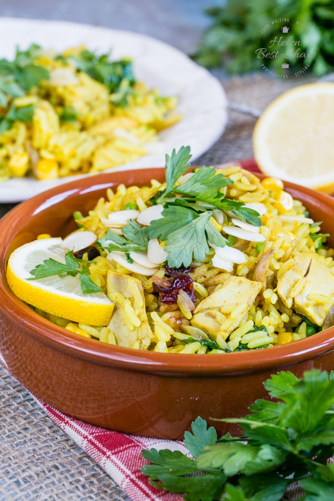 A closeup of leftover roast turkey biryani. The vibrant yellow turkey and rice dish is in a shallow brown bowl, garnished with a slice of lemon, slivered almonds and coriander leaves. In the background is a white plate on which more curry has been served.