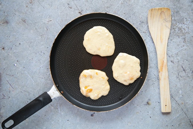 A top down view of a frying pan with three mango pancakes. There are bubbles showing on the top, indicating that the pancakes are ready to flip.