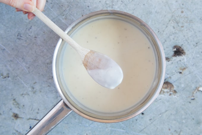 A saucepan of white sauce. A wooden spoon is held above the sauce. The back of the spoon is coated with the white sauce.