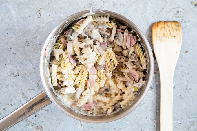 A picture from above of a saucepan full of cooked fusilli pasta, turkey, ham, mushrooms, onion and sauce, all mixed together. Next to the saucepan is a wooden spatula.