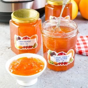 A picture of two pots of Instant Pot Seville orange marmalade. The nearer pot has been opened, and a spoon is in the jar. In the foreground is a small dish filled with more marmalade.