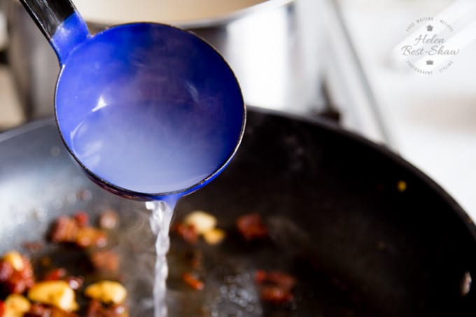 Adding pasta water to the sauce results in a silky smooth sauce that evenly coats the pasta. This picture shows a deep blue ladle of cloudy pasta water being poured into a pan of pasta sauce that's in the background of the shot, out of focus.