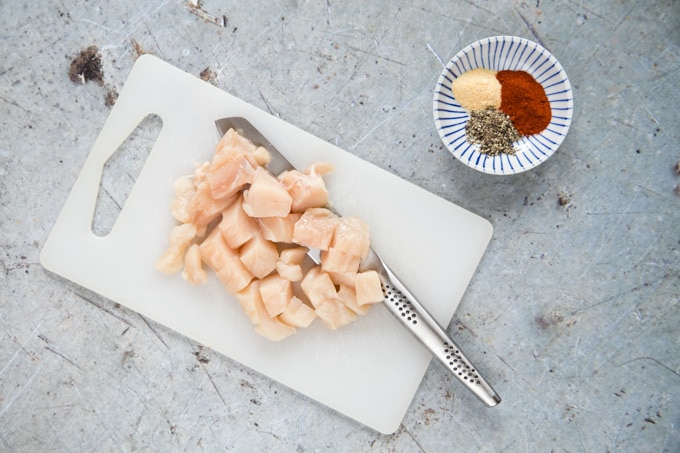 A top down view of a chopping board holding a raw chicken breast that has been cut into cubes. Next to the board is a small dish holding black pepper, garlic granules, and paprika.