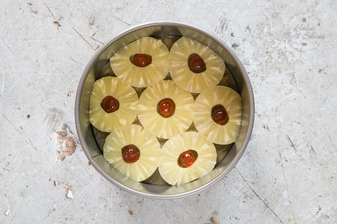 A top down view of a cake tin containing 7 pineapple ringe, each with a glace cherry in the middle, arrainged in a circle around a central ring. 